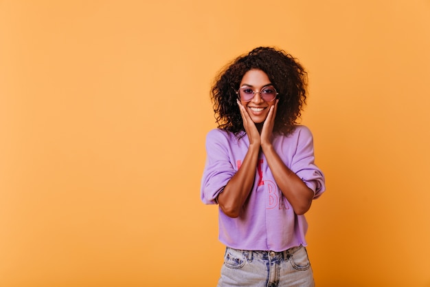 Satisfeita garota negra em óculos de sol da moda, se divertindo durante o ensaio fotográfico interno. Mulher africana elegante refinada divertidamente sorrindo em amarelo.