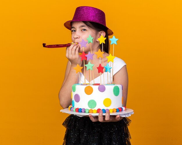 Foto grátis satisfeita jovem caucasiana com chapéu de festa roxo segurando um bolo de aniversário e soprando o apito de festa isolado na parede laranja com espaço de cópia