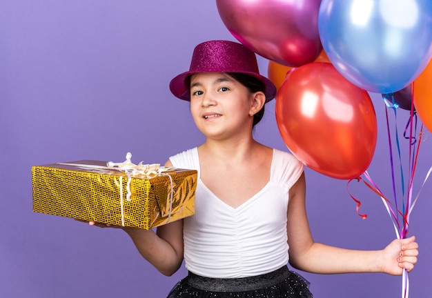 Satisfeita jovem caucasiana com chapéu de festa violeta segurando balões de hélio e uma caixa de presente isolada na parede roxa com espaço de cópia