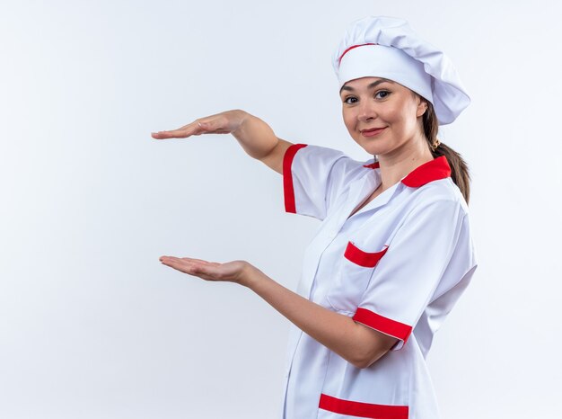 satisfeita jovem cozinheira vestindo uniforme de chef fingindo estar segurando algo isolado na parede branca