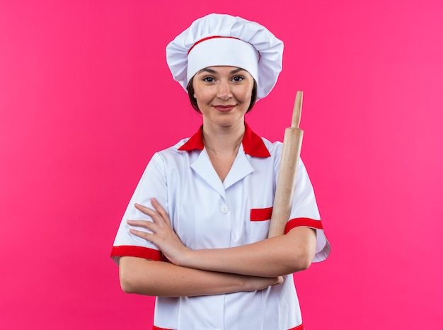 Foto grátis satisfeita jovem cozinheira vestindo uniforme de chef segurando o rolo de massa cruzando as mãos isoladas na parede rosa