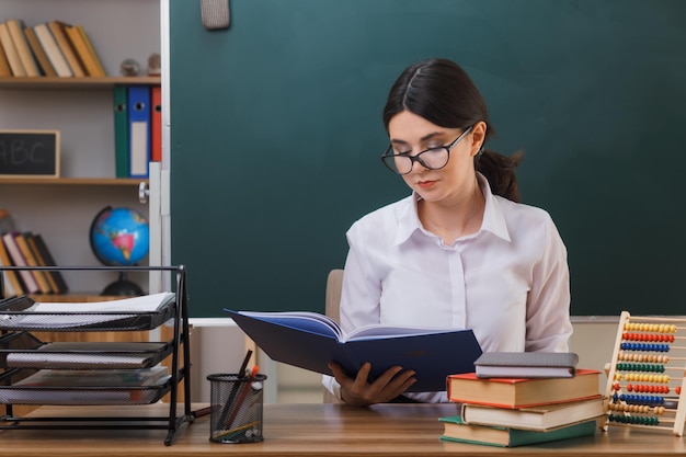 Satisfeita jovem professora usando óculos segurando e olhando para o livro sentado na mesa com ferramentas escolares em sala de aula