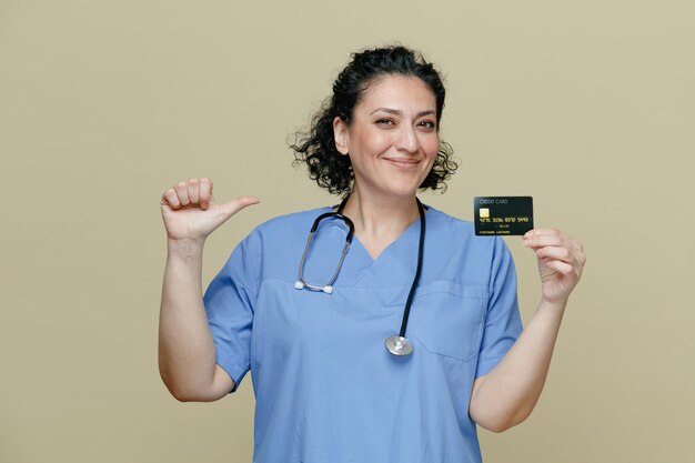 Satisfeita médica de meia idade vestindo uniforme e estetoscópio no pescoço, mostrando o cartão de crédito olhando para a câmera apontando para si mesma isolada no fundo verde-oliva