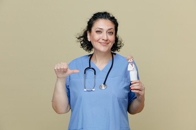 Satisfeita médica de meia-idade vestindo uniforme e estetoscópio no pescoço segurando estatueta de médico apontando para ele olhando para câmera isolada em fundo verde-oliva