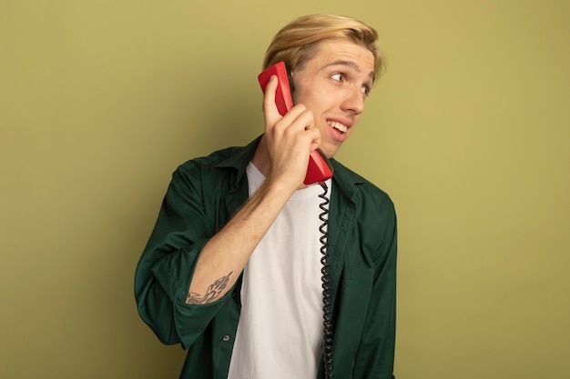 Satisfeito ao olhar para o lado, jovem loiro de camiseta verde fala ao telefone