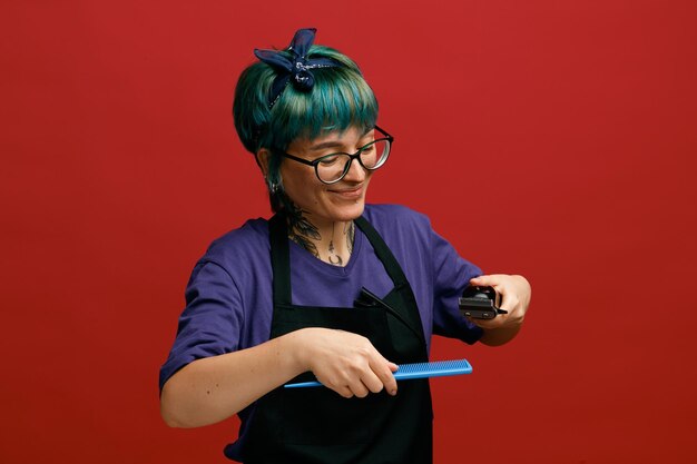 Foto grátis satisfeito jovem barbeiro feminino usando óculos uniformes e bandana segurando pente e aparador de cabelo no ar olhando para baixo fingir fazer corte de cabelo para cliente isolado em fundo vermelho