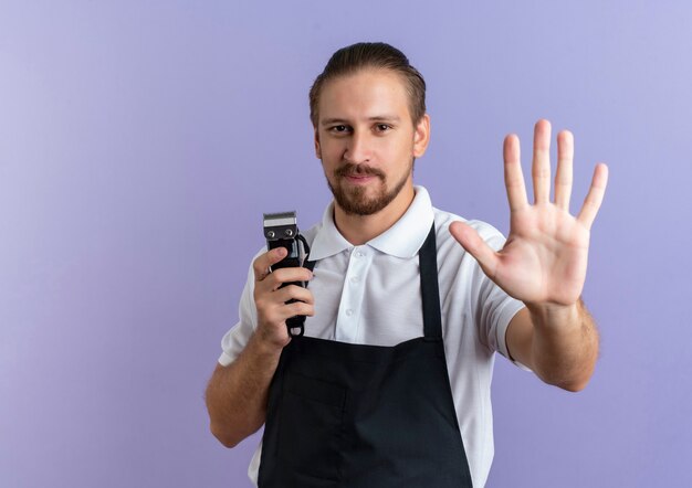 Satisfeito, jovem e bonito barbeiro vestindo uniforme, segurando uma tesoura de cabelo e estendendo a mão para a frente, isolado na parede roxa