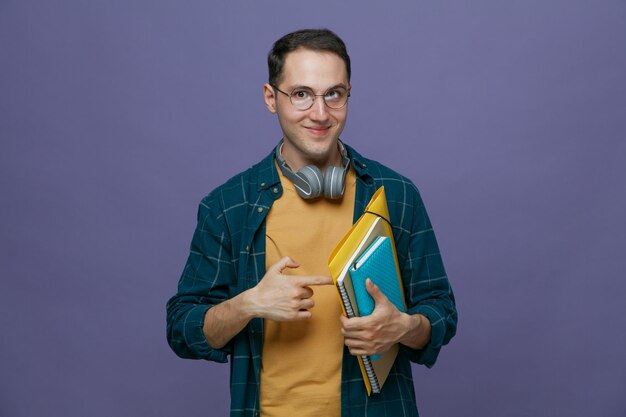 Satisfeito jovem estudante do sexo masculino usando fones de ouvido de óculos no pescoço segurando o bloco de notas da pasta caderno apontando para eles olhando para a câmera isolada no fundo roxo