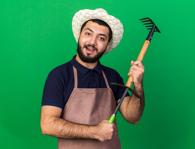 satisfeito jovem jardineiro caucasiano com chapéu de jardinagem, segurando o ancinho sobre o ancinho de enxada isolado na parede verde com espaço de cópia