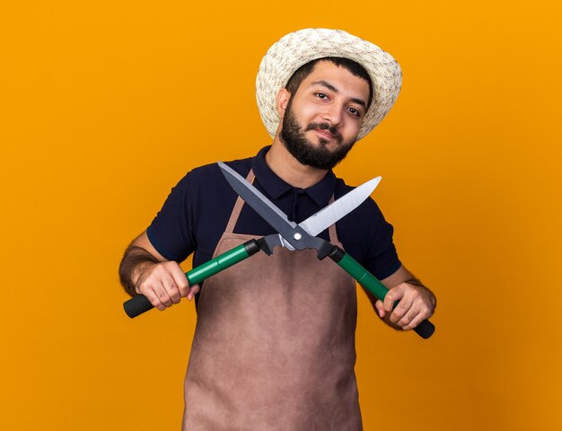 satisfeito jovem jardineiro caucasiano usando chapéu de jardinagem segurando uma tesoura de jardinagem isolada na parede laranja com espaço de cópia