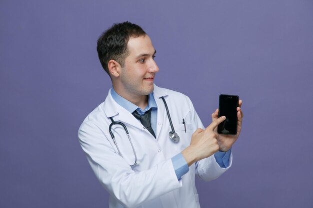 Satisfeito jovem médico masculino vestindo roupão médico e estetoscópio no pescoço mostrando o celular olhando para ele apontando o dedo sobre ele isolado no fundo roxo