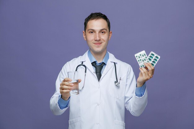 Satisfeito jovem médico masculino vestindo roupão médico e estetoscópio no pescoço, olhando para a câmera segurando o copo de água mostrando pacotes de comprimidos isolados no fundo roxo