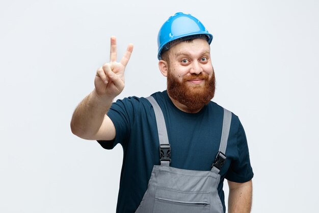 Satisfeito jovem trabalhador da construção civil usando capacete de segurança e uniforme olhando para câmera mostrando sinal de paz isolado no fundo branco