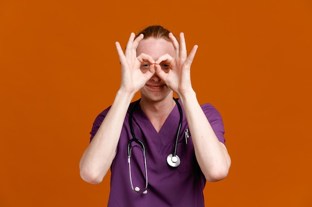 Foto grátis satisfeito mostrando gesto de máscara jovem médico masculino vestindo uniforme com estetoscópio isolado em fundo laranja