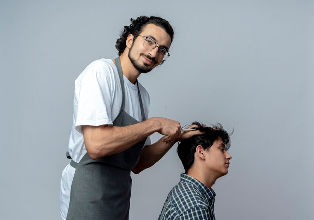 Satisfeito o jovem barbeiro caucasiano usando óculos e faixa de cabelo ondulado em uniforme em pé em vista de perfil fazendo corte de cabelo para seu jovem cliente, isolado no fundo branco com espaço de cópia