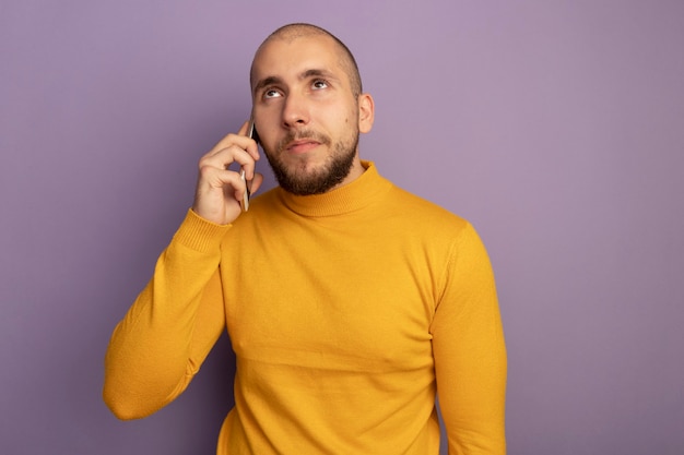 Satisfeito olhando para cima, jovem bonito falando no telefone isolado na parede roxa