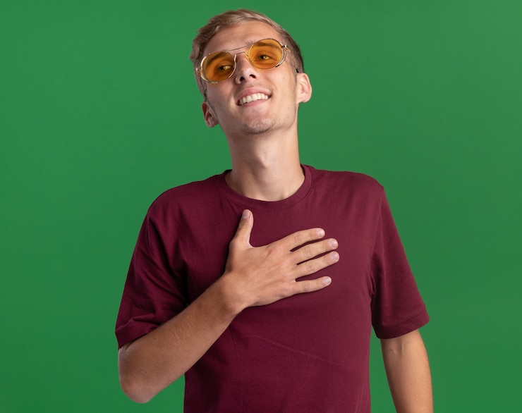 Foto grátis satisfeito, olhando para o lado, jovem bonito, vestindo camisa vermelha e óculos, colocando a mão no coração isolado na parede verde