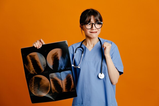 Satisfeito segurando a jovem médica de raio-x usando estetoscópio uniforme de quinto isolado em fundo laranja