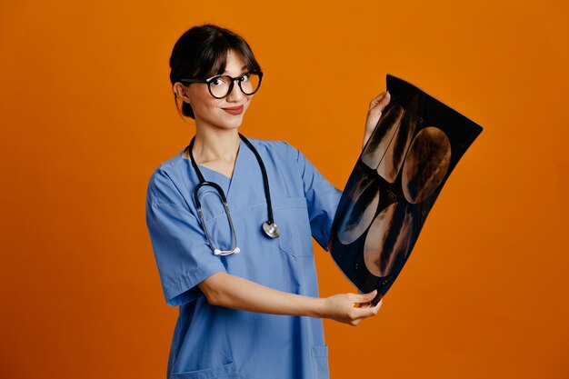 Satisfeito segurando a jovem médica de raio-x usando estetoscópio uniforme de quinto isolado em fundo laranja