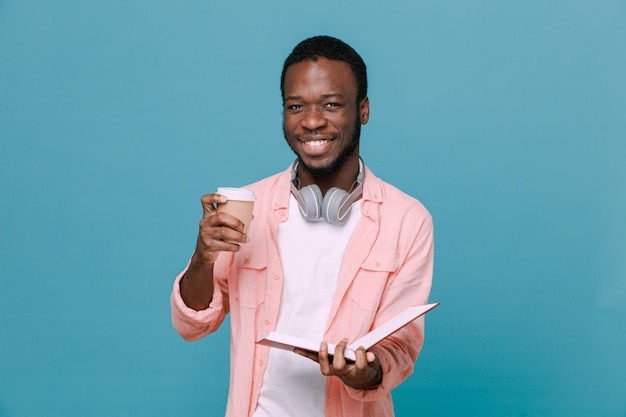 Satisfeito segurando a xícara de café com livro jovem afro-americano usando fones de ouvido no pescoço isolado em fundo azul