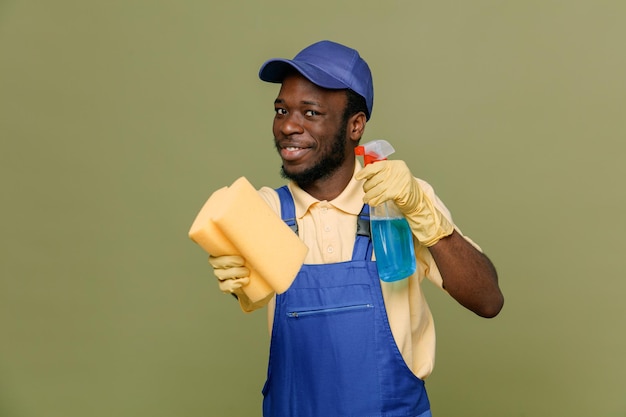 Satisfeito segurando o agente de limpeza com esponja jovem limpador afro-americano de uniforme com luvas isoladas em fundo verde