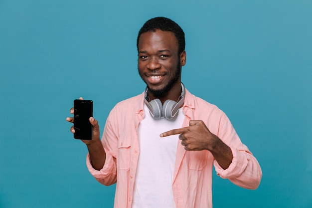 Foto grátis satisfeito segurando o telefone jovem afro-americano usando fones de ouvido no pescoço isolado em fundo azul