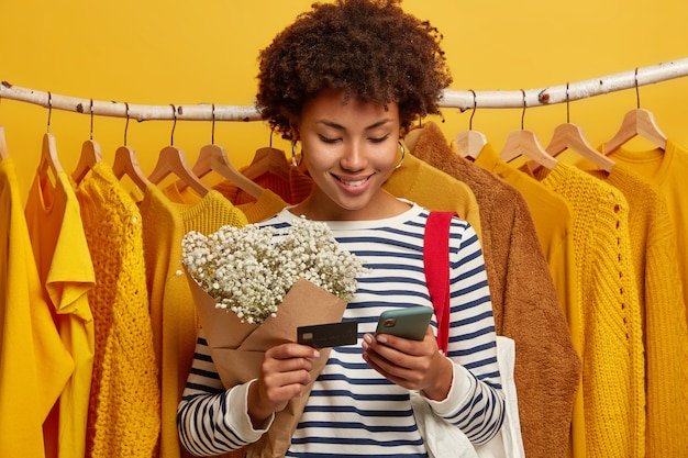Satisfeito shopaholic fica perto de uma variedade de roupas em cabides, compra roupas online ou paga pela compra com cartão de creadit e aplicativo para celular