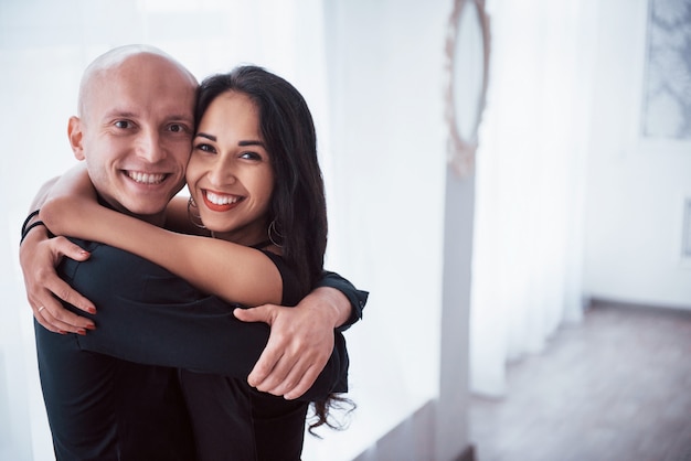 Se abraçam e sorriem. Retrato de casal feliz dentro de casa. Cara careca e mulher morena fica na sala branca