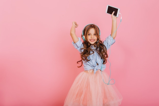 Foto grátis se divertindo, expressando emoções positivas verdadeiras de uma jovem incrível alegre ouvindo música através de fones de ouvido isolados no fundo rosa. infância feliz de criança fofa. lugar para texto