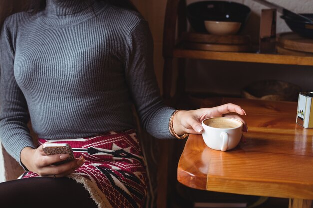 Seção intermediária da mulher usando telefone celular enquanto toma uma xícara de café