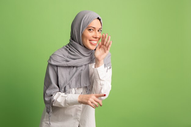 Segredo, conceito de fofoca. Mulher árabe feliz em hijab. Retrato de menina sorridente, posando no fundo verde do estúdio.