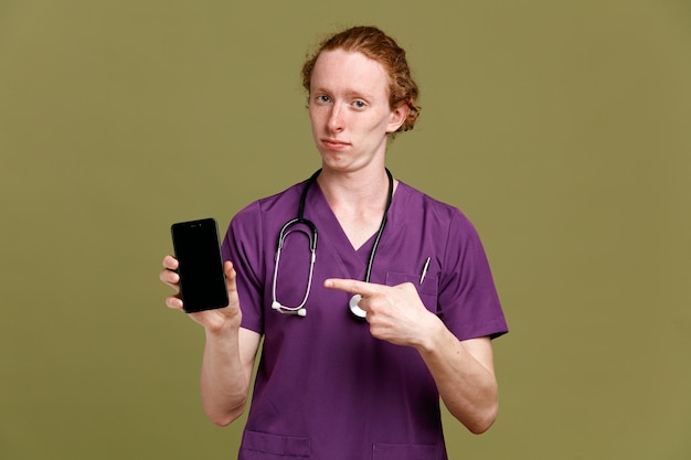 Segurando confiante e aponta para telefone jovem médico masculino vestindo uniforme com estetoscópio isolado em fundo verde