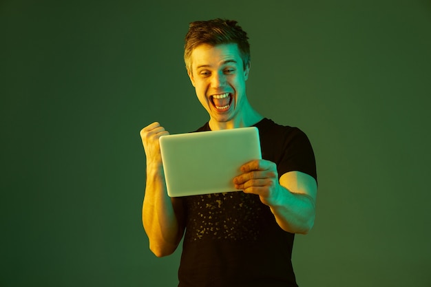 Foto grátis segurando o tablet, comemorando a vitória na aposta ou no jogo. retrato do homem caucasiano sobre fundo verde do estúdio em luz de néon. lindo modelo masculino. conceito de emoções humanas, expressão facial, vendas, anúncio.