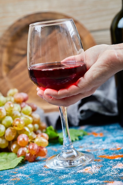 Segurando um copo de vinho tinto em uma mesa azul com um cacho de uvas