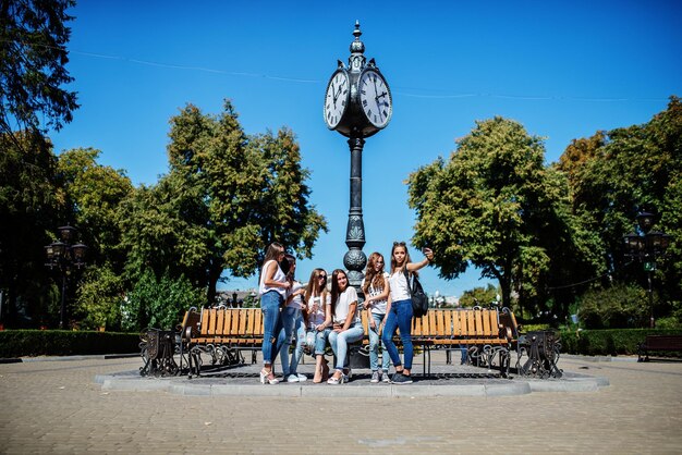 Seis lindas garotas sentadas em um banco ao lado do velho relógio de rua no parque