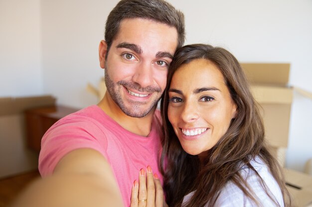 Selfie de um jovem casal feliz em sua nova casa, posando com caixas de papelão no fundo, segurando o gadget na mão