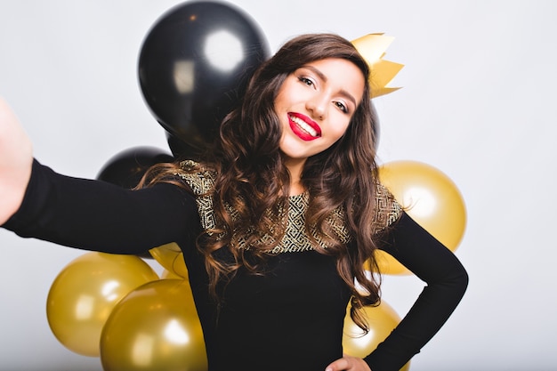 Selfie retrato alegre mulher com cabelo longo encaracolado morena, coroa amarela, vestido preto de luxo. Comemorando ano novo, festa de aniversário, se divertindo com balões dourados e pretos.