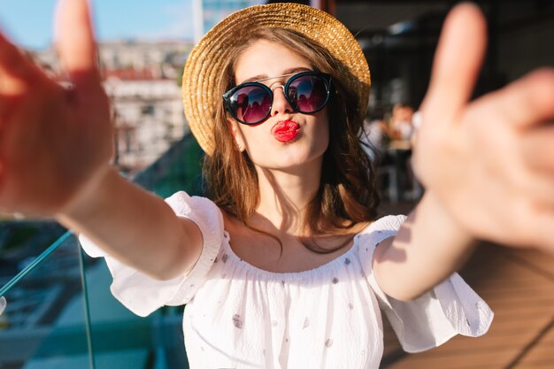 Selfie-retrato de close-up de uma garota engraçada com cabelo comprido em pé na luz do sol no terraço. Ela usa vestido branco, chapéu, batom vermelho, óculos escuros. Ela manda um beijo para a câmera.