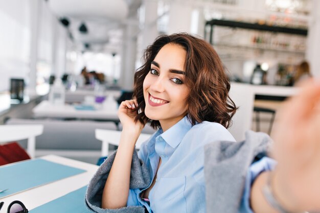 Selfie-retrato de uma linda garota morena com cabelo curto, sentado à mesa em manta cinza no terraço no restaurante. Ela usa camisa azul e parece feliz.