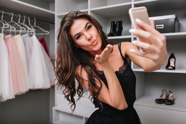 Selfie-retrato de uma linda menina morena fazendo uma selfie usando um smartphone em seu camarim. Ela manda um beijo. Suas roupas elegantes, expressando verdadeiras emoções faciais positivas.