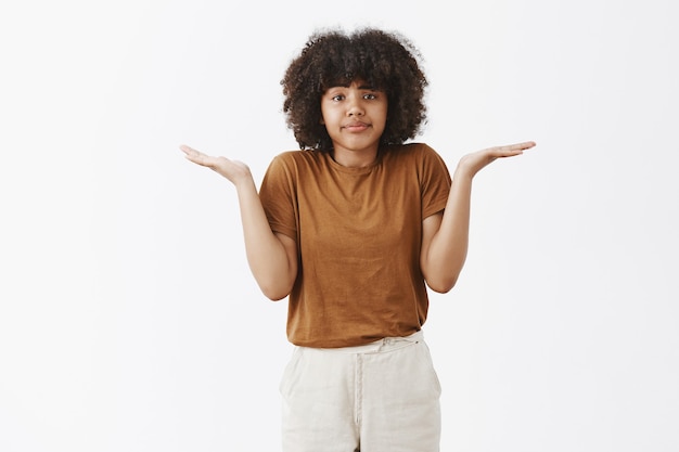 Sem noção, atraente e elegante mulher urbana jovem com penteado afro em uma camiseta marrom dando de ombros com as mãos abertas franzindo os lábios sem interesse e sem ideia