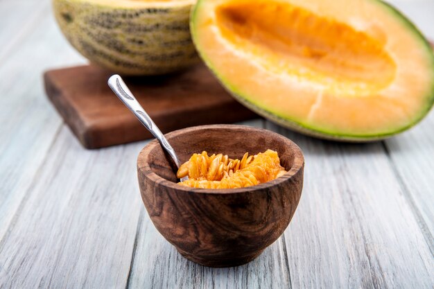 Sementes amarelas de melão cantalupo em uma tigela de madeira com fatias de melão na superfície de madeira cinza