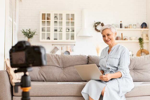 Sempre em contato. Na moda, elegante mulher autônoma sênior vestida de azul, sentada no sofá com o computador portátil no colo, gravando vídeo para blog, contando segredos de negócios, tendo um olhar animado