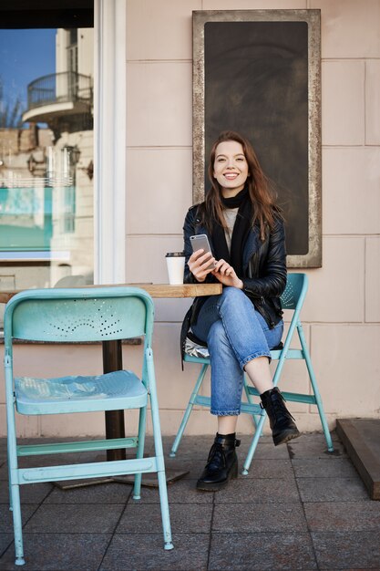 Sempre há tempo para o café. Retrato de mulher bonita feliz sentado no pátio do café, segurando o smartphone e olhando