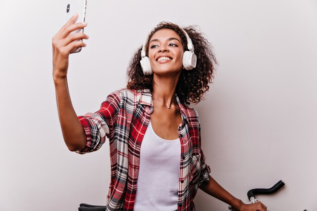 Senhora africana inspirada em fones de ouvido brancos, tirando foto de si mesma. Modelo feminino interessado em camisa quadriculada fazendo selfie com expressão de rosto feliz.