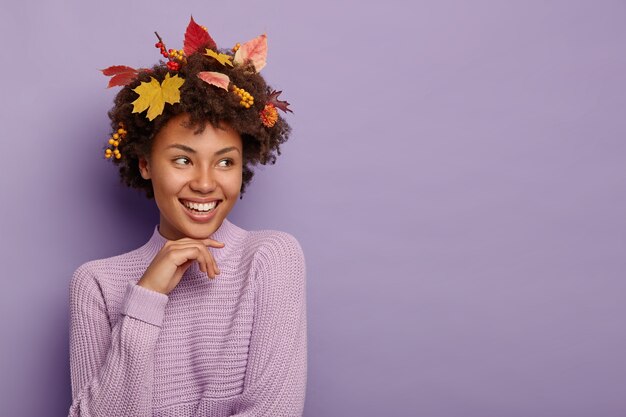 Senhora afro-americana positiva com folhas de outono no cabelo encaracolado e frutos maduros, usa um suéter roxo de malha, focado à parte, isolado sobre a parede violeta, copie a área