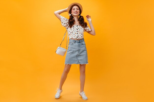 Senhora alegre vestida com saia jeans e top recortado posando com bolsa branca em fundo laranja.