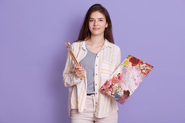 Senhora artista de pé contra a parede do estúdio lilás, adorável jovem segurando uma nova foto e pincel nas mãos