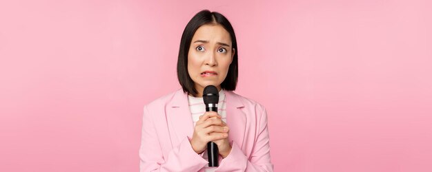 Senhora asiática ansiosa de terno falando em público dando discurso com microfone na conferência parecendo assustada em pé sobre fundo rosa