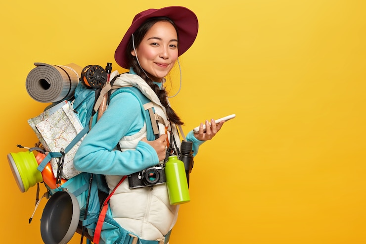 Foto grátis senhora asiática com expressão satisfeita, tenta encontrar rota com mapa de navegação online, segura telefone celular, usa chapéu, roupas casuais, carrega mochila, frasco, binóculos, isolado na parede amarela